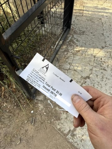 Hand holding a receipt showing "Total Paid: $5.00" and "Issued: 0915" near a metal gate on a concrete surface.
