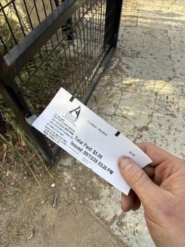 A hand holds a receipt displaying "Total Paid: $5.00", "1 Adult, Resident", and "Issued: 09/15/20 05:25 PM" in front of a metal gate on a paved surface.