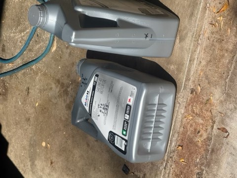 Two gray motor oil containers on concrete ground with blue hose attached to one.