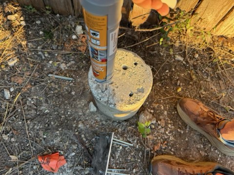 Hand holds tube inserted into concrete block on ground with dirt, rocks, small plant, and shoe nearby.