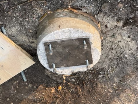 Concrete cylinder with metal plate secured by four bolts, resting on dirt ground with a wooden plank nearby.