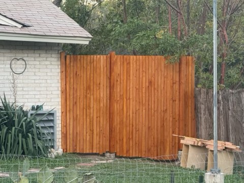 Light brown wooden fence adjacent to white brick house with heart-shaped cutout, green plants, wire mesh fence, and trees.