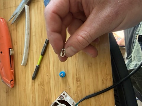 Hand holding small white component over bamboo surface with tools including orange utility knife, green screwdriver, and blue screw.