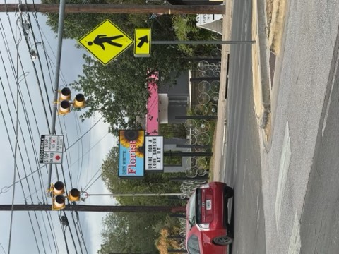 Street scene with pedestrian crossing sign, traffic lights, and a red car near a sidewalk.