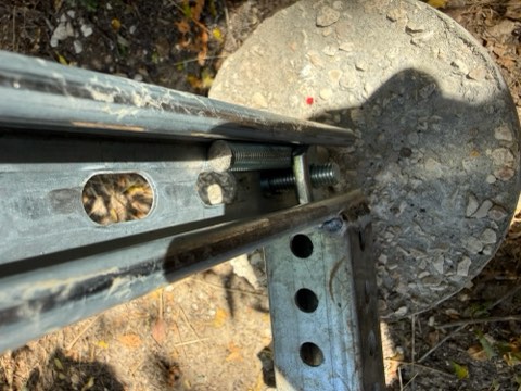 Close-up of metal fence components with holes, bolts, and a concrete disc on ground with leaves.
