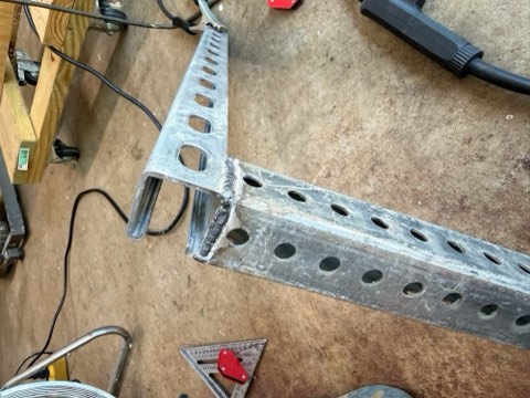 Metal C-channel with multiple holes and welded joint on concrete floor, surrounded by tools like angle ruler, power tool, and wooden workbench.
