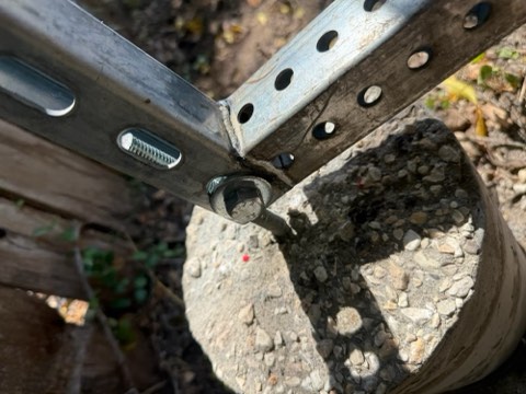 Metal fence structure with welded joints and concrete base, showing bolted connections and gravel-filled concrete.