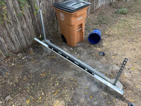 Metal frame structure with concrete bases, brown trash bin, and blue bucket on dirt ground near wooden fence.