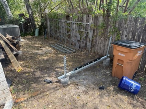Backyard scene with wooden fence, trash bin, blue Lowe bucket, and metal brackets on concrete bases.